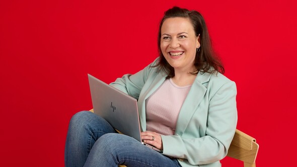 Unsere Kollegin sitzt auf einem Stuhl Unsere Kollegin sitzt auf einem Holzstuhl vor einem roten Hintergrund. Ihre Beine hängen entspannt über die Stuhllehne. Sie hat einen Laptop auf dem Schoß und lächelt freundlich in die Kamera.