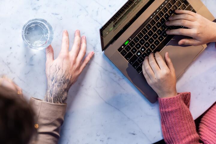 Arbeitssituation bei OTTO Zwei Kollegen sitzen nebeneinander am Tisch. Auf dem Tisch steht ein Glas Wasser und ein geöffneter Laptop. Einer von ihnen tippt auf dem Laptop. Zu sehen sind nur die Hände der Kollegen.