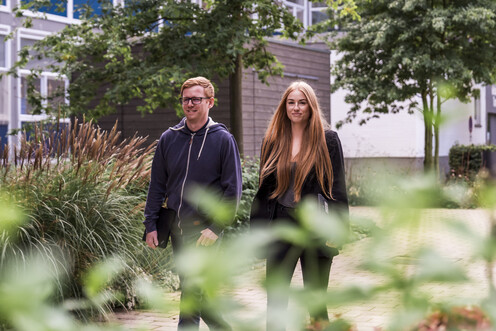 Der Campus Boulevard Zwei Mitarbeitende gehen auf dem gepflasterten Weg des Campus Boulevards entlang. An der Seite des Weges sind viele Grünflächen mit Büschen und Bäumen angelegt und im Hintergrund steht ein Gebäude