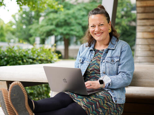 Expertin Technical Writing sitzt auf einer Holzbank Eine Expertin Technical sitzt lächelnd mit ihrem Laptop auf einer Holzbank auf dem Otto Campus.