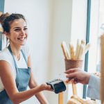 ine Person im Verkauf hält einer kaufenden Person das Kartenlesegerät entgegen. Diese hält die Karte nur in die Nähe.