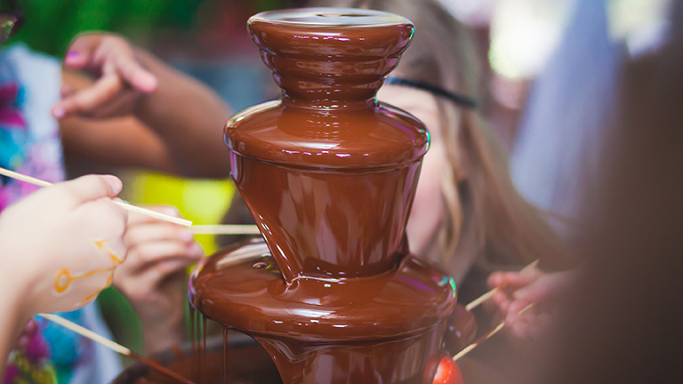Mehrere Kinder sitzen um einen Schokobrunnen und halten Früchte in die Schokolade.