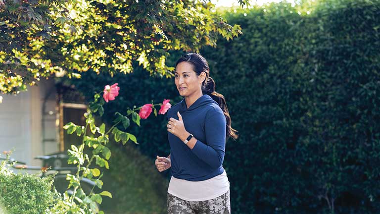 Eine Frau joggt durch eine gartenähnliche Kulisse. Sie trägt einen blauen Kapuzenpullover und am Handgelenk das Fitness-Armband Inspire 2 von Fitbit.