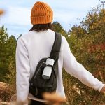 Ein Mann streift durch ein Feld. An seinem Rucksack hängt der Bluetooth-Lautsprecher Sonos Roam in weiß.