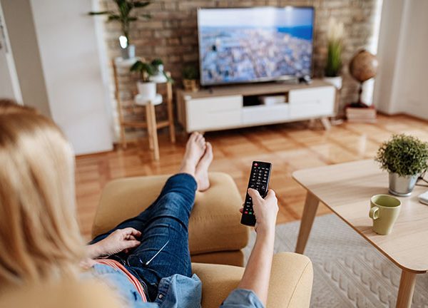 Eine Person sitzt auf einem Sessel mit einer Fernbedienung in der Hand und schaut Fernsehen.