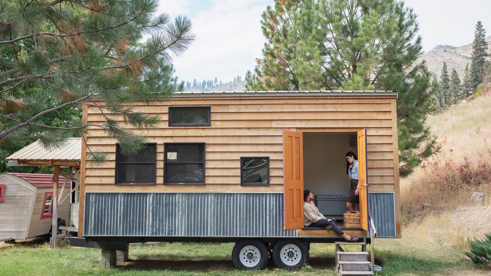 Zwei Personen sitzen am Eingang eines Tiny House, das auf einen Anhänger gebaut ist.