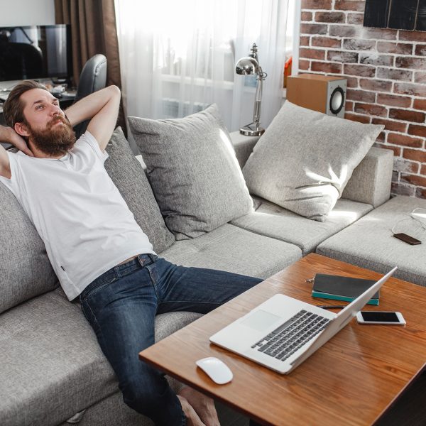 Eine Person sitzt auf einem Sofa. Vor ihr auf dem Tisch ist ein MacBook zu sehen, neben dem Sofa ein Lautsprecher.