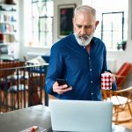 Eine Person steht mit einer Tasse in der Hand vor einem Laptop. In der Hand hält sie ein iPhone.