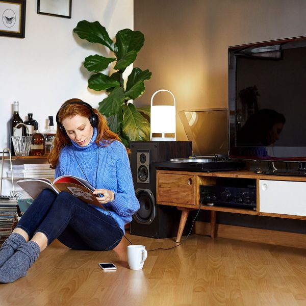 Eine Frau sitzt auf dem Fußboden und liest in einem Magazin, hinter ihr sind ein Sideboard mit TV-Gerät und Lautsprechern rechts und links zu sehen.