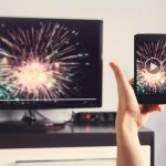 Eine Hand hält ein Smartphone vor einem Smart-TV, auf beiden Geräten ist das gleiche Bild zu sehen, ein Feuerwerk.