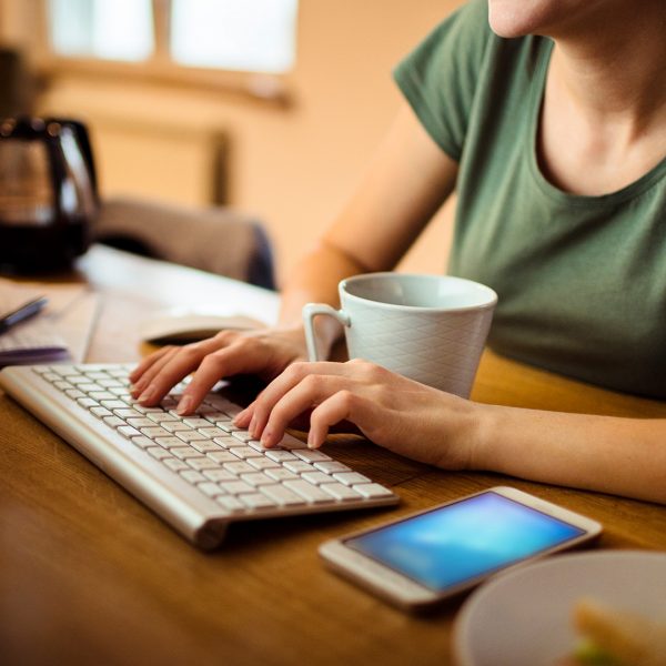 Eine Person sitzt vor einem Monitor und tippt auf einer Tastatur. Vor ihr liegt ein Smartphone.