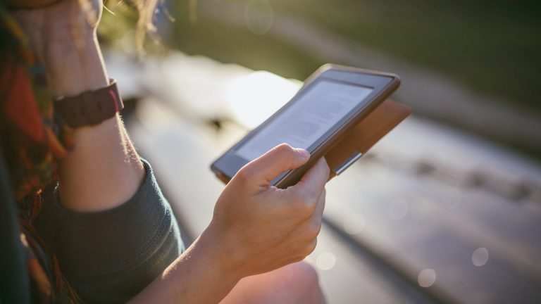 Eine Person sitzt im Freien und hält einen E-Book-Reader in der Hand.