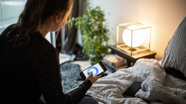 Eine Person sitzt auf einem Bett und steuert per Smartphone eine Lampe vor ihr.