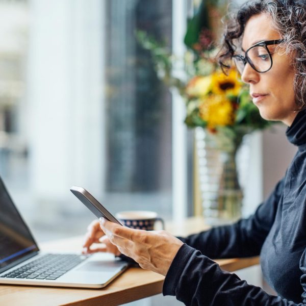 Eine Person sitzt vor einem Laptop. In der Hand hält sie ein Smartphone, auf das sie schaut.