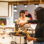 Eine Person steht vor einem Slow-Cooker-Kochtopf und rührt darin mit einem Holzlöffel.