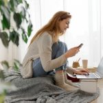 Eine Person sitzt auf einem Sitzkissen vor einem Laptop. In der Hand hält sie ein Smartphone.