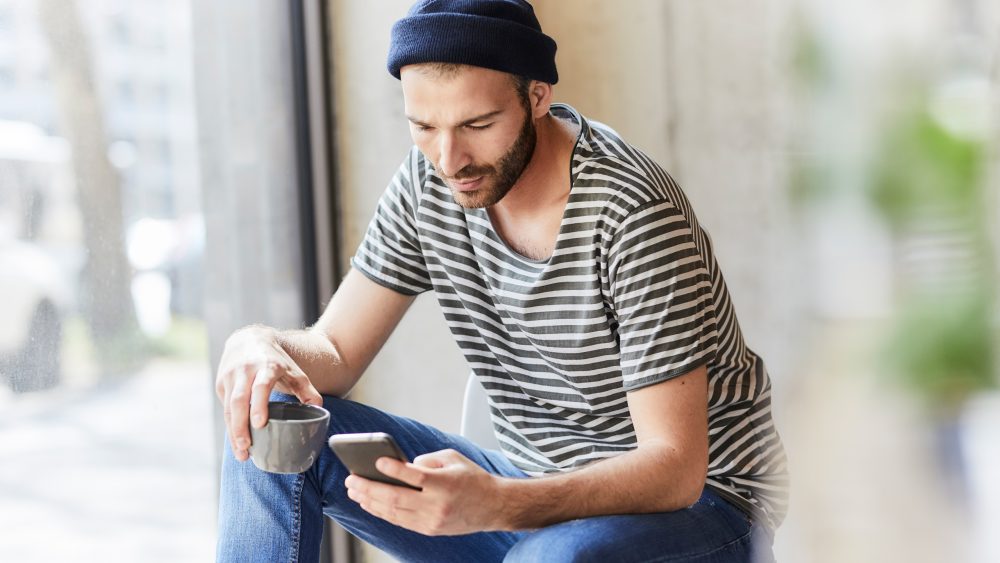 Eine Person mit Kaffeetasse in der Hand schaut auf das Smartphone in der anderen Hand.