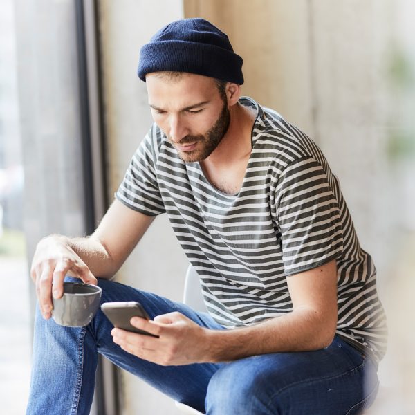 Eine Person mit Kaffeetasse in der Hand schaut auf das Smartphone in der anderen Hand.