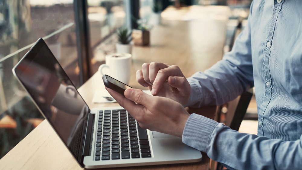 Eine Person sitzt in einem Café vor einem MacBook und bedient derweil ein Smartphone in der Hand.