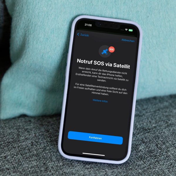 Ein iPhone 14 lehnt auf einem Sofa an einem Kissen. Auf dem Bildschirm ist der Startscreen zum Notruf SOS über Satellit zu sehen.