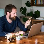 Eine Person sitzt an einem Tisch vor dem Laptop und macht etwas auf ihrem Smartphone.