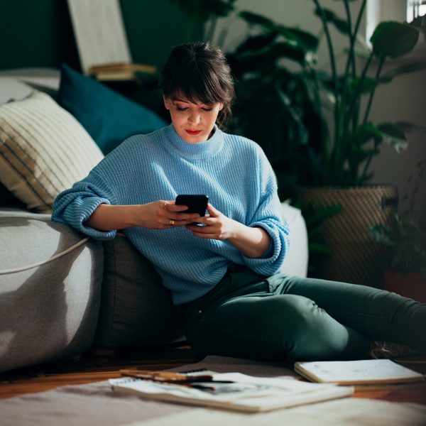 Eine Person sitzt vor einer Couch auf dem Boden und schaut auf ihr Smartphone.