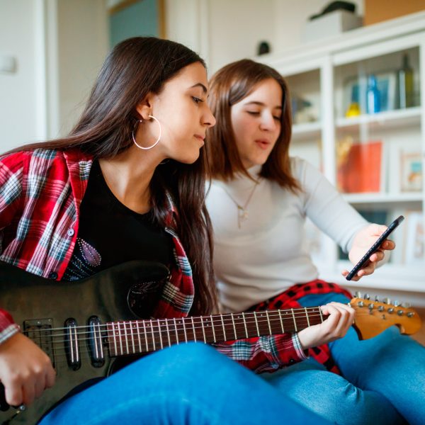 Zwei Personen sitzen auf dem Boden. Eine hält eine E-Gitarre in der Hand, die andere ein Smartphone hoch, auf dem wohl Noten zu sehen sind.
