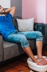 Ein Mensch sitzt mit hochgekrempelter Jeans auf einer grauen Couch. Die Füße sind in einem weiß-rosé-farbenen und mit Wasser gefüllten Fußmassagegerät.