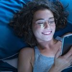 Eine Person mit braunen Locken und silberner Brille liegt in einem Bett mit blauer Bettwäsche und schaut auf ihr Smartphone.