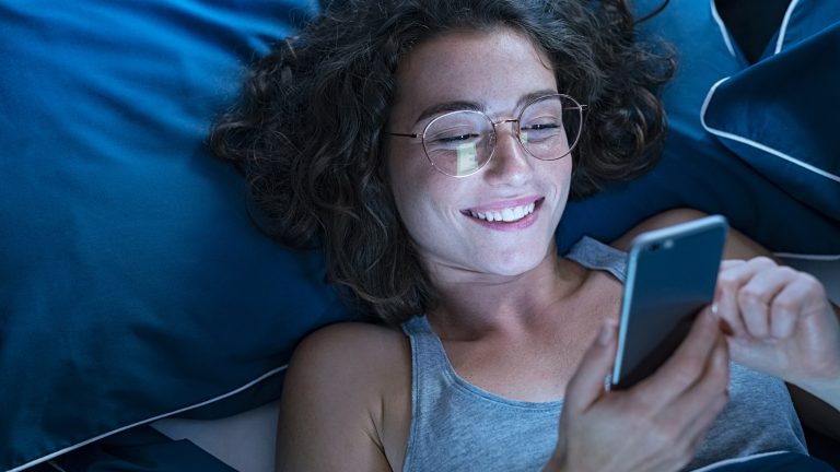 Eine Person mit braunen Locken und silberner Brille liegt in einem Bett mit blauer Bettwäsche und schaut auf ihr Smartphone.