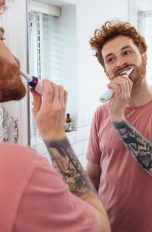Eine Person steht in einem Badezimmer vor einem Spiegel und putzt sich die Zähne mit einer elektrischen Zahnbürste.