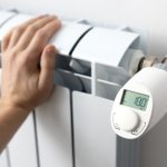 Eine Person fasst an eine Heizung, an der ein elektronisches Thermostat sitzt.