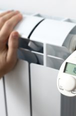 Eine Person fasst an eine Heizung, an der ein elektronisches Thermostat sitzt.