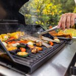 Eine Person steht mit einer Grillzange an einem Gasgrill und wendet Fisch.