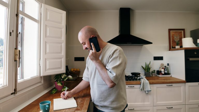 Eine Person steht in der Küche, telefoniert mit ihrem Handy und schreibt in ein Notizbuch.