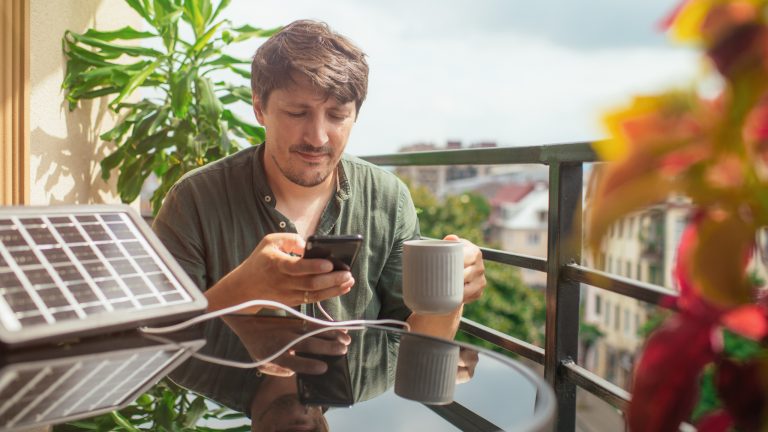 Ein Mensch sitzt an einem Balkontisch, auf dem ein mittelgroßes Solarladegerät steht, an das sein Smartphone angeschlossen ist.
