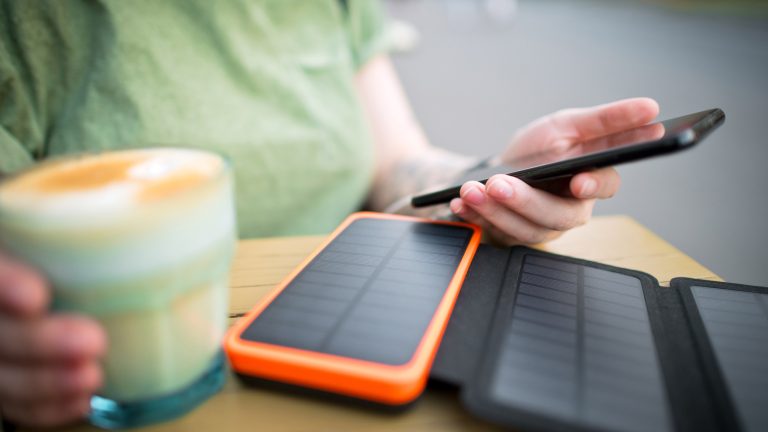 Eine Person sitzt mit einem Kaffee an einem Holztisch, auf dem ein ausklappbares Solar-Ladegerät liegt, das mit ihrem Handy verbunden ist.