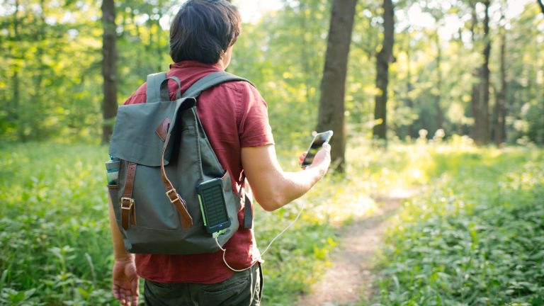 Eine Person geht im Wald spazieren und hat eine Solar-Powerbank außen an ihren Rucksack geklemmt und mit ihrem Smartphone verbunden.