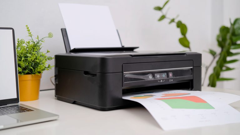 Ein Drucker steht neben einem Laptop und einer Pflanze auf einem Tisch und druckt ein Blatt mit bunten Mustern.