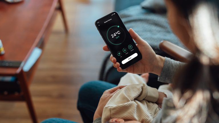 Eine Person sitzt mit ihrem Baby auf der Couch und stellt über ihr Smartphone die Temperatur der Klimaanlage ein.