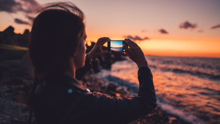 Eine Person steht an einem Stand und fotografiert das Meer bei Sonnenuntergang.