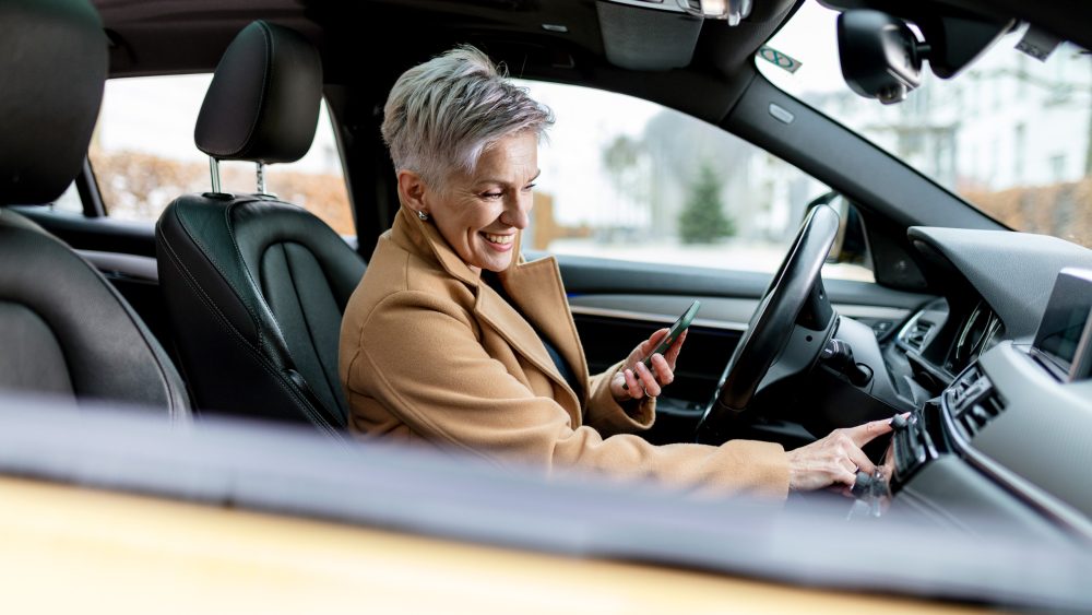 Eine Person sitzt in einem Auto und tippt auf dem Infotainment-System des Autos herum. In der anderen Hand hält sie ein Smartphone.