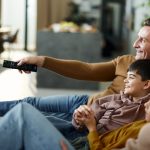 Zwei Personen und zwei Kinder sitzen nebeneinander auf der Couch im Wohnbereich. Eine Person richtet eine Fernbedienung auf etwas in der Ferne.