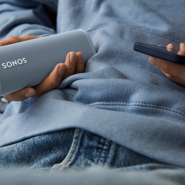 Eine Person hält einen Sonos-Lautsprecher in der einen, ein Smartphone in der anderen Hand.