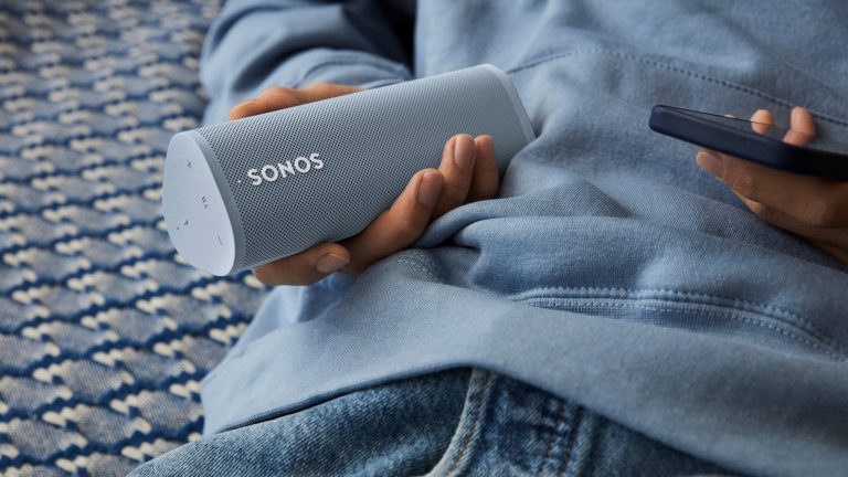 Eine Person hält einen Sonos-Lautsprecher in der einen, ein Smartphone in der anderen Hand.