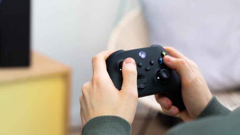 Eine Person hält den Controller einer Xbox Series X in der Hand. Die Konsole selbst ist unscharf im Hintergrund zu erkennen.