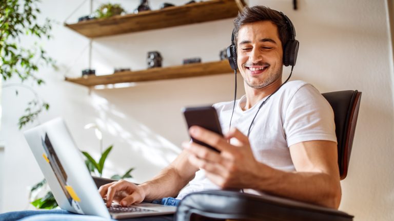 Eine Person sitzt auf einem Sessel, trägt Kopfhörer auf dem Kopf und bedient einen Laptop und ein Smartphone gleichzeitig.