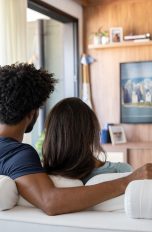 Ein Pärchen sitzt im Wohnzimmer auf der Couch und sieht auf einem Fernseher fern. 