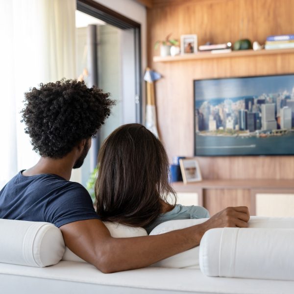 Ein Pärchen sitzt im Wohnzimmer auf der Couch und sieht auf einem Fernseher fern. 