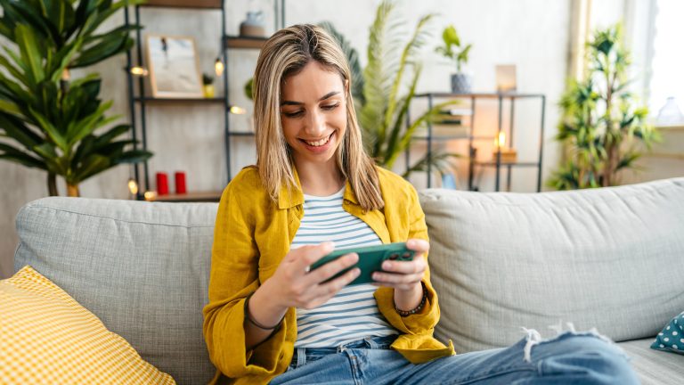 Eine Person sitzt auf einem Sofa und schaut lächelnd auf einem Smartphone etwas an.